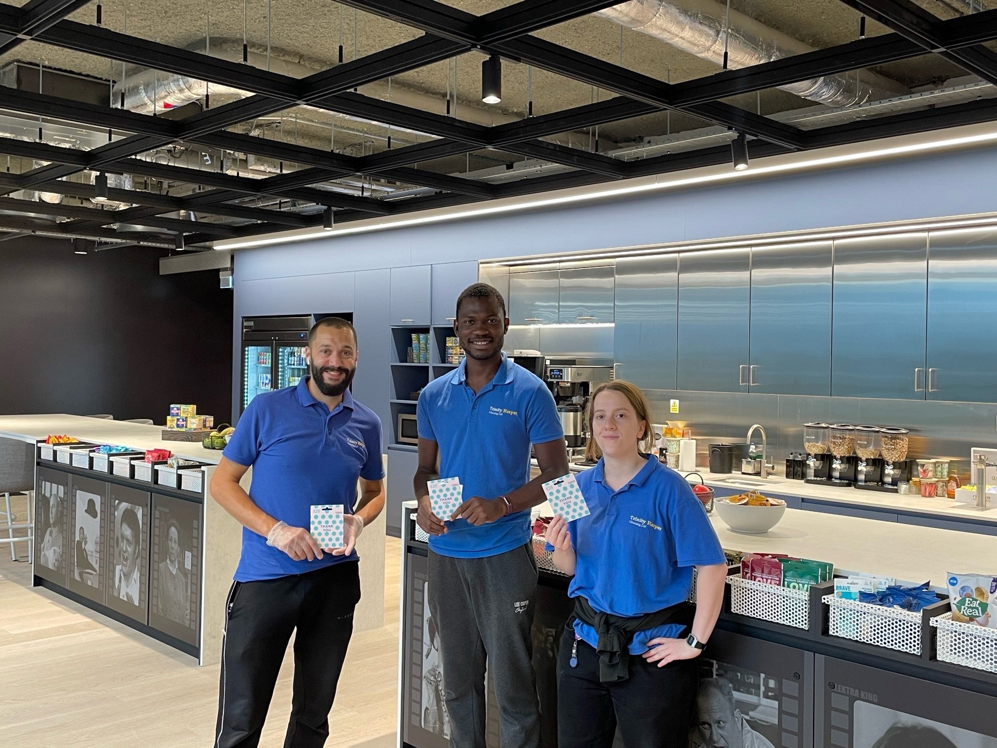 3 people in blue shirts stood in a clean cafeteria holding gift cards