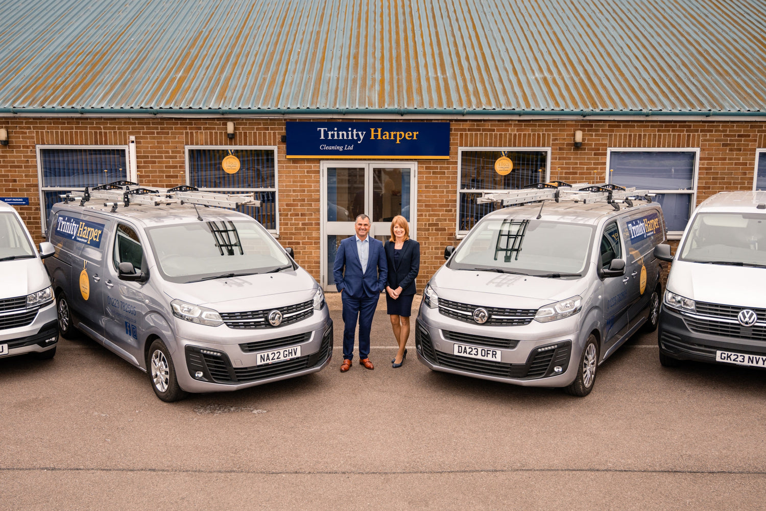Two people standing in front of four vans outside a building with 'Trinity Harper' branding.